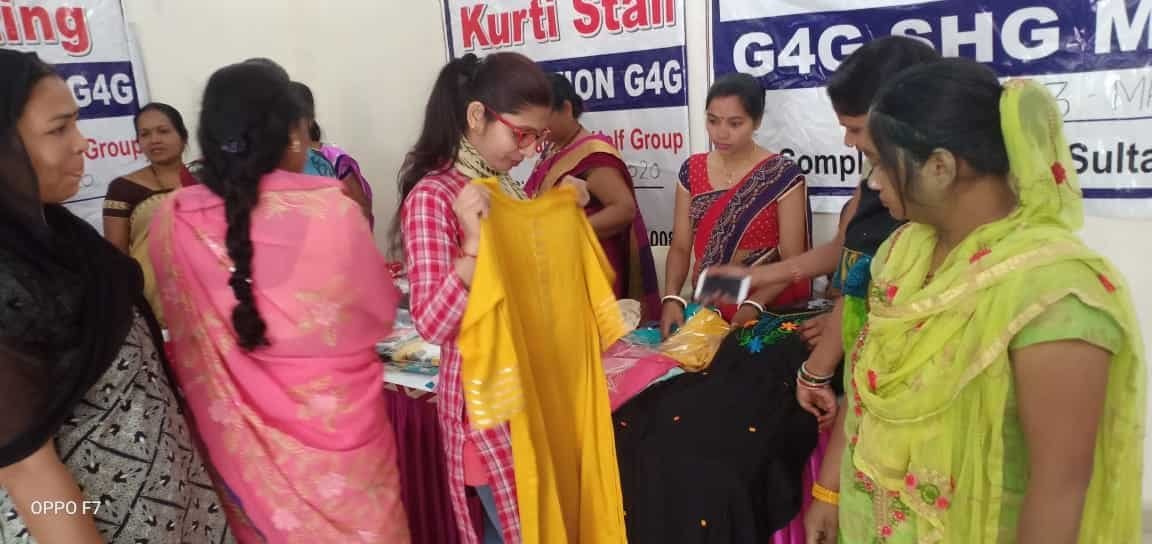 Women examining a yellow garment and a black embroidered garment at a G4G stall.
